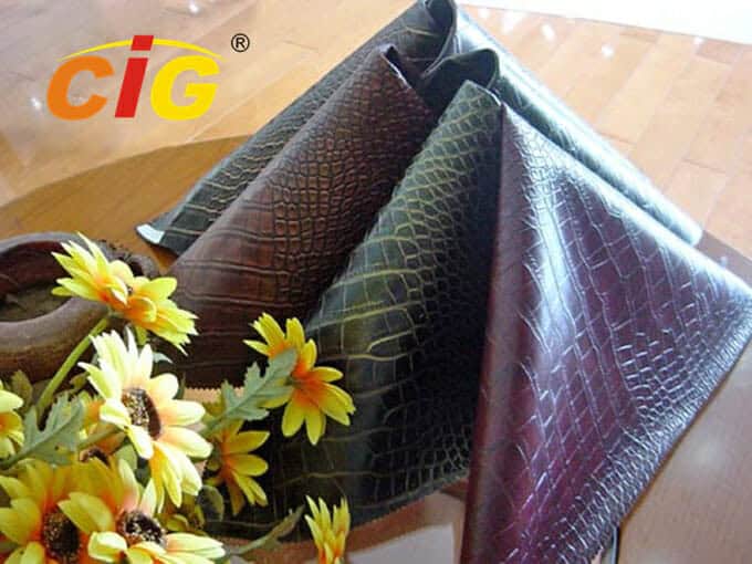Eine Ausstellung von Stoffmustern aus künstlichem Krokodilleder, elegant drapiert neben einer Schale mit gelben Blumen auf einem Holztisch.
